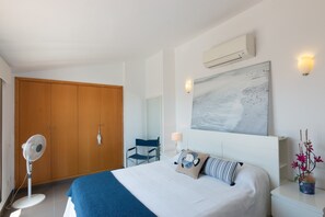 A hotel room with a bed, a standing fan, a wooden wardrobe, a chair, a wall-mounted air conditioner, and a painting. - Apartment 'Cala Gaviota' with Sea Views, Wi-Fi, and Air Conditioning (Colònia de Sant Jordi)