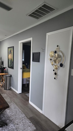 Una habitación con pared gris, un reloj decorativo, una puerta y un vistazo a una cama al fondo.