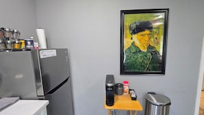 Una cocina con refrigerador, cafetera, basurero y un cuadro en la pared.