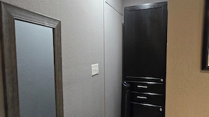 Un pasillo con un espejo, una puerta negra, un panel de interruptores blanco y un pequeño estante con una caja.