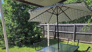 Un patio al aire libre con una mesa de vidrio y sillas metálicas, un área techada con un paraguas estampado y una cerca de madera al fondo.