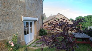 Un bâtiment en pierre avec une porte blanche, un petit jardin avec de l’herbe et une plante en pot, et une antenne parabolique sur le toit.