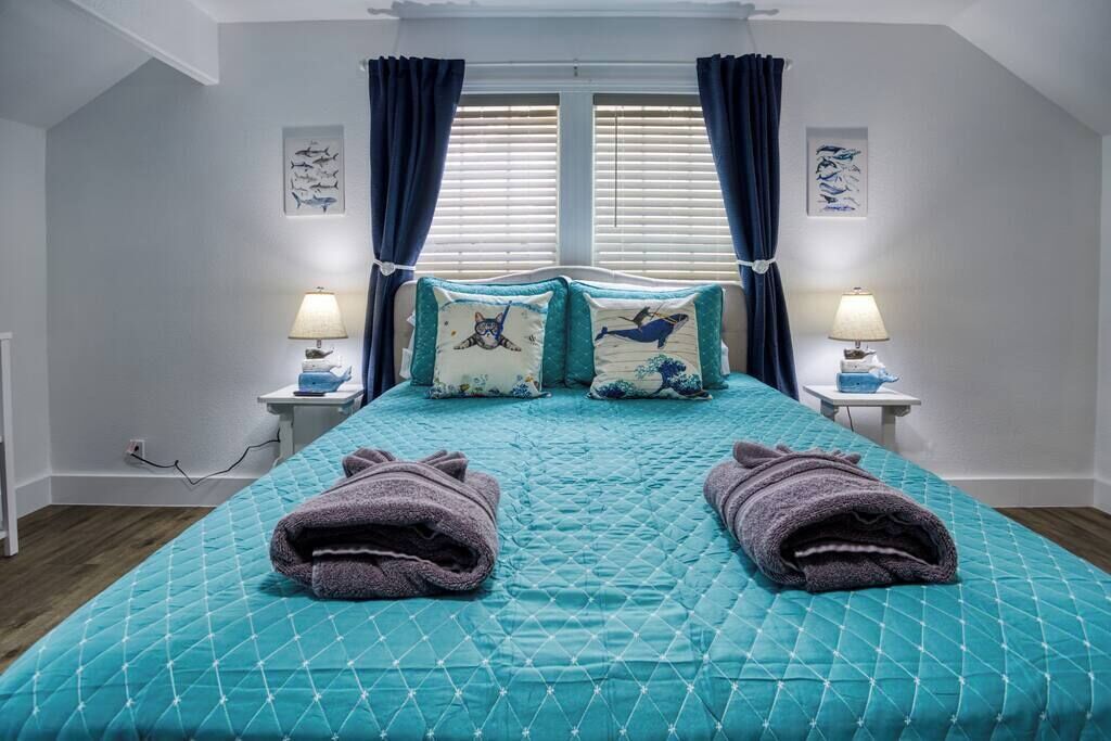 Un dormitorio con colcha acolchada color aguamarina, dos almohadas con motivos marinos y dos lámparas de noche sobre mesitas blancas.