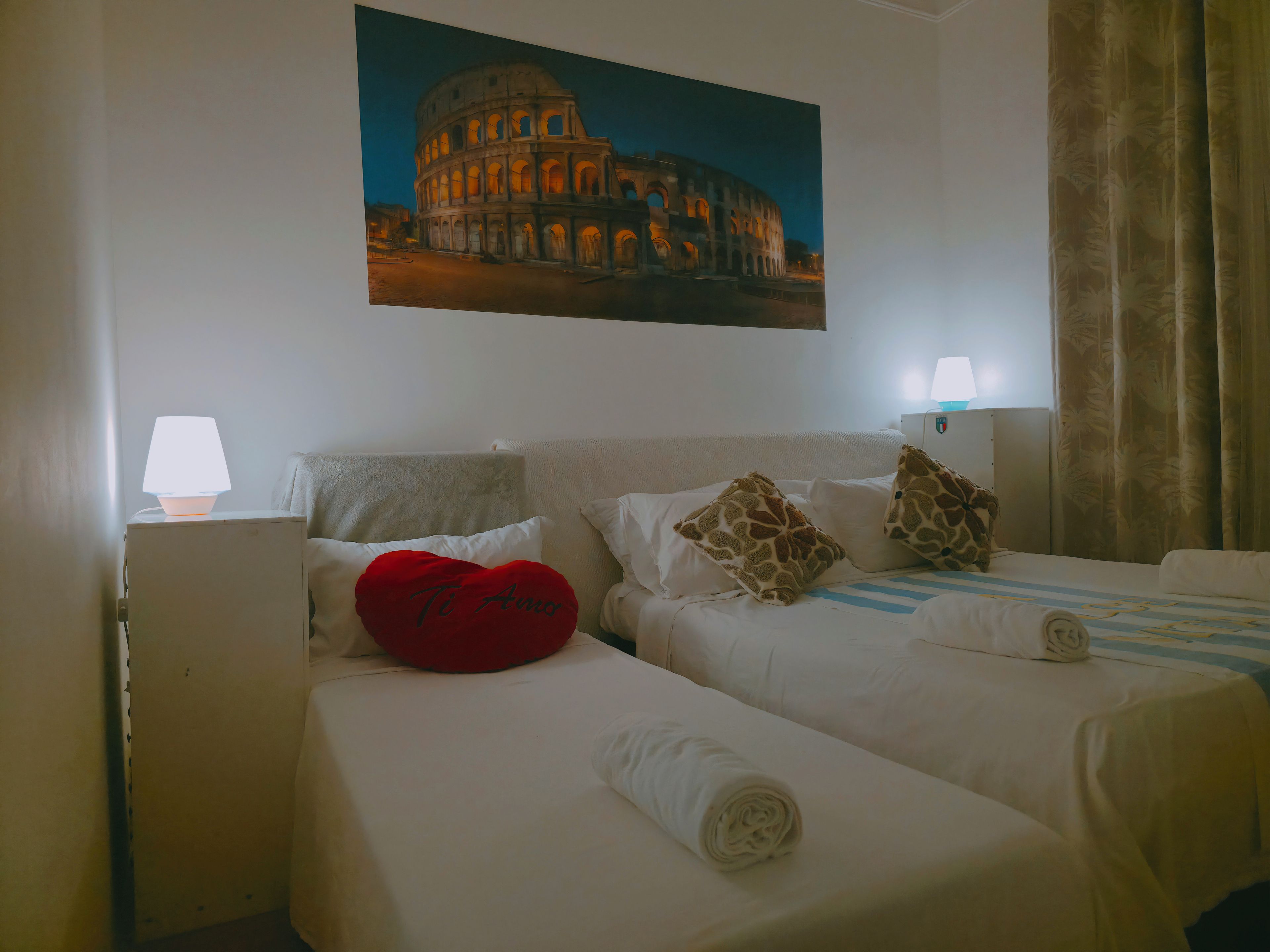A hotel room with two beds, a painting of the Colosseum, a red heart-shaped pillow, and a lamp on the nightstand.