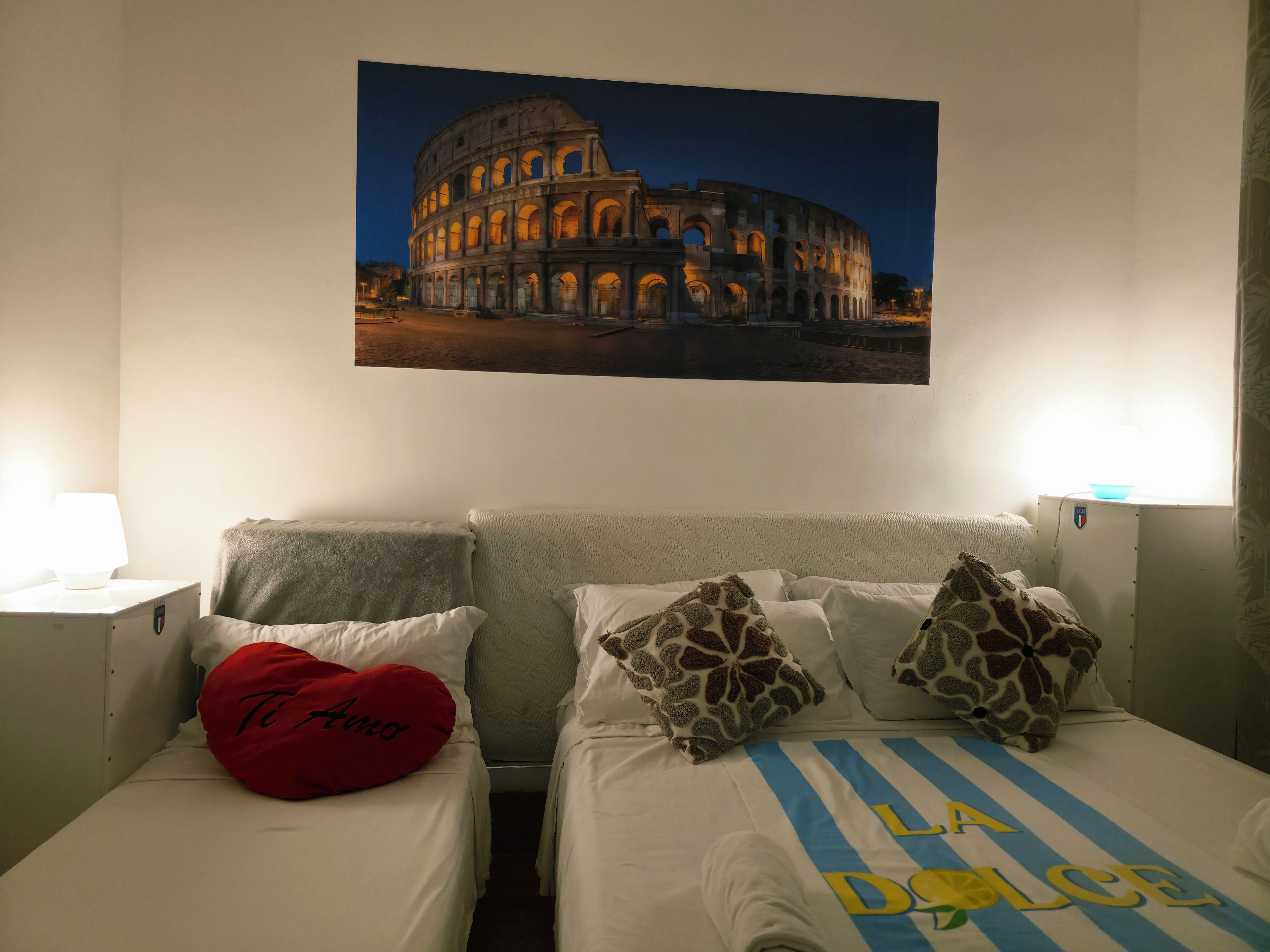 Habitación de hotel con dos camas, un cojín rojo con forma de corazón y un cuadro colgado en la pared del Coliseo.