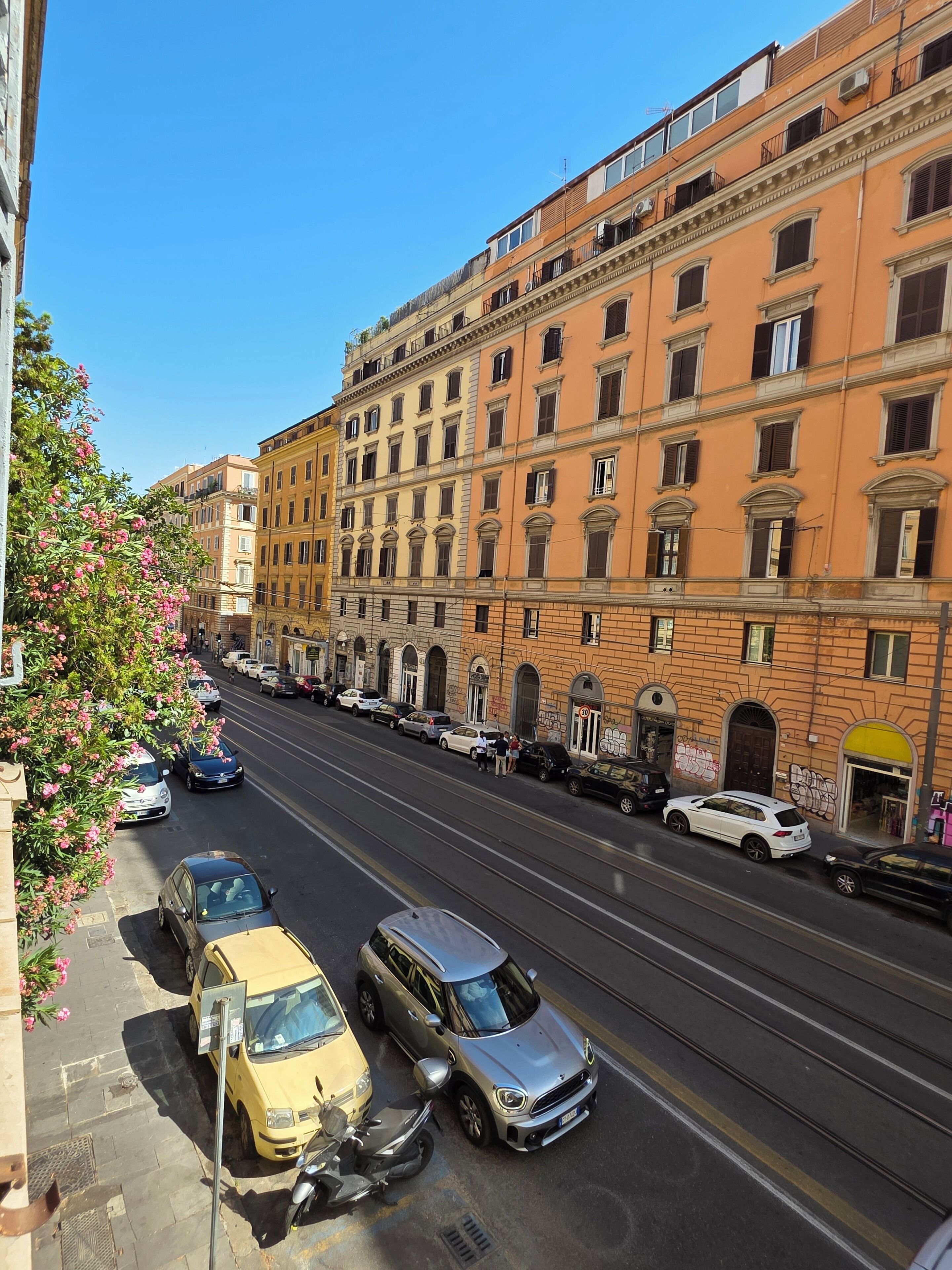 Un immeuble à plusieurs étages, avec une rangée de voitures garées et une ligne de tramway qui longe la rue.