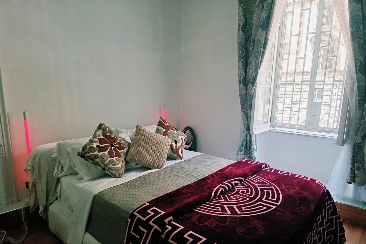 A bedroom with a bed, pillows, a blanket, a window with curtains, and a bedside lamp.