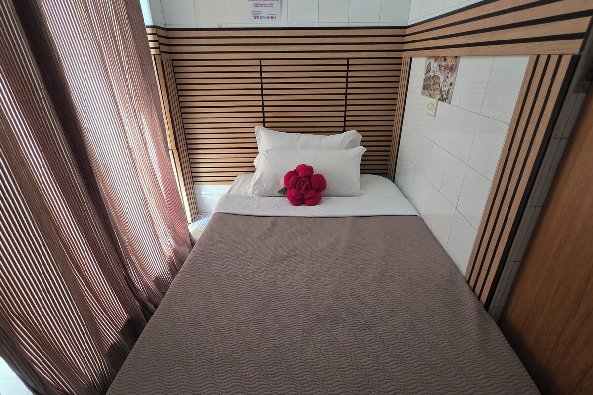 A hotel room with a bed, a wooden headboard, a white wall with a poster, and a window with blinds.