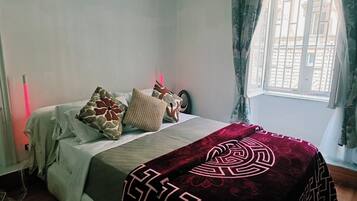 Un dormitorio con cama, almohadas, manta, ventana con cortinas y una lámpara de noche.