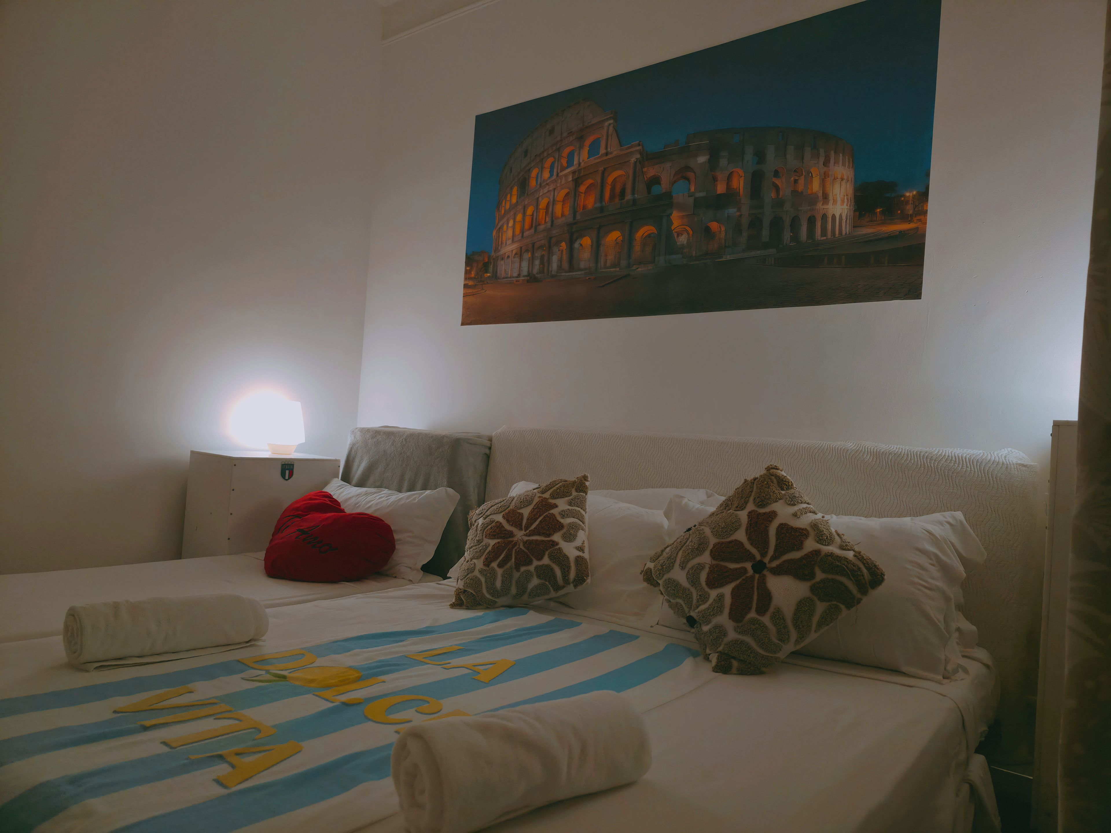 A hotel room with a bed, pillows, a lamp, and a painting of the Colosseum.