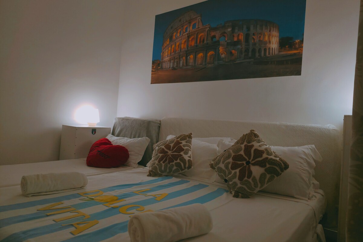 A hotel room with a bed, pillows, a lamp, and a painting of the Colosseum.