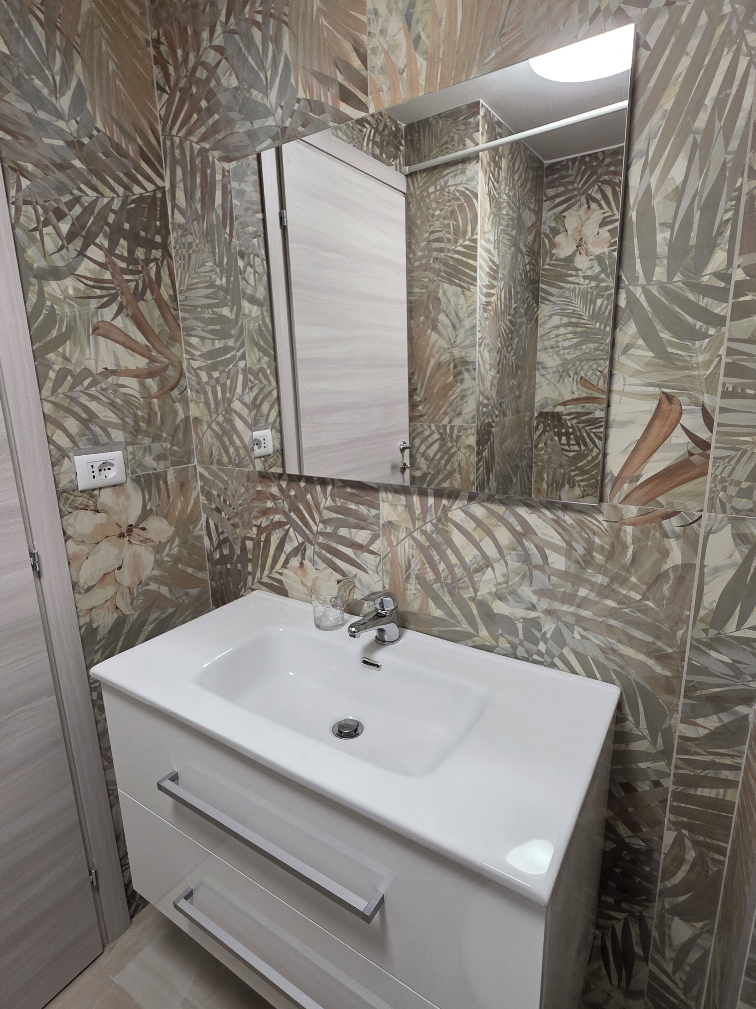 Une salle de bain avec un lavabo blanc, un miroir au-dessus et un mur orné d’un motif de feuilles.