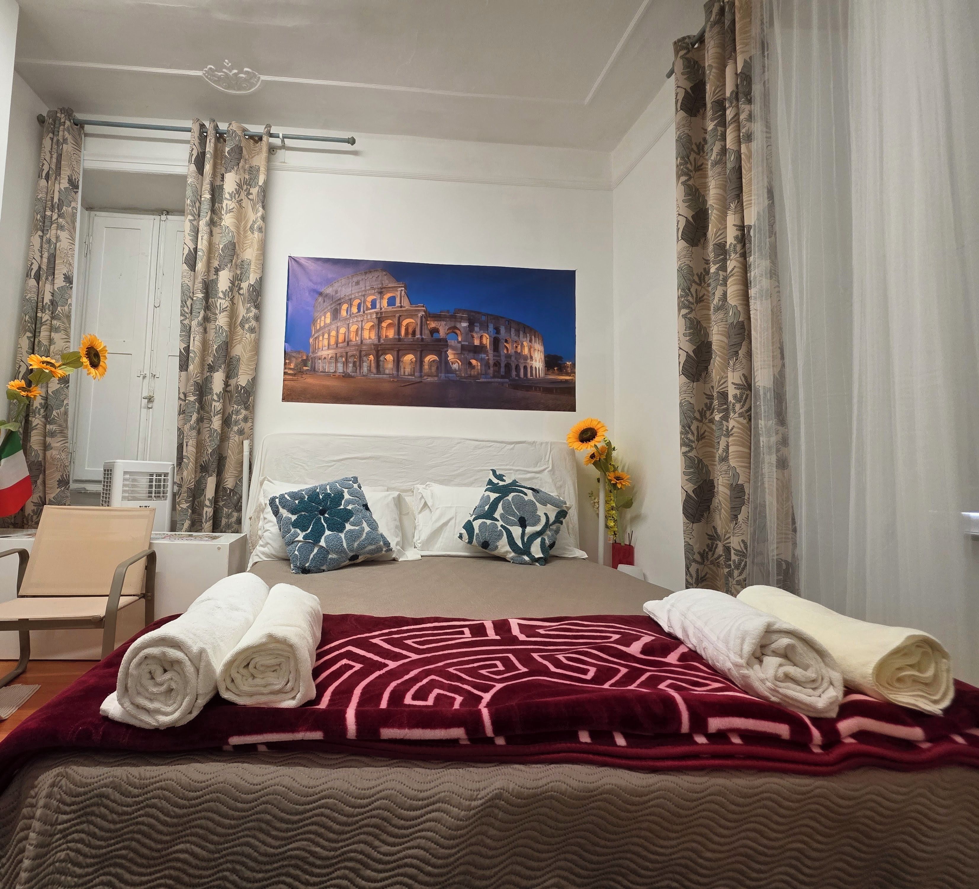 Hotellihuone, jossa on sänky, tyynyjä, peitto, kuva Colosseumista, työpöytä ja tuoli.