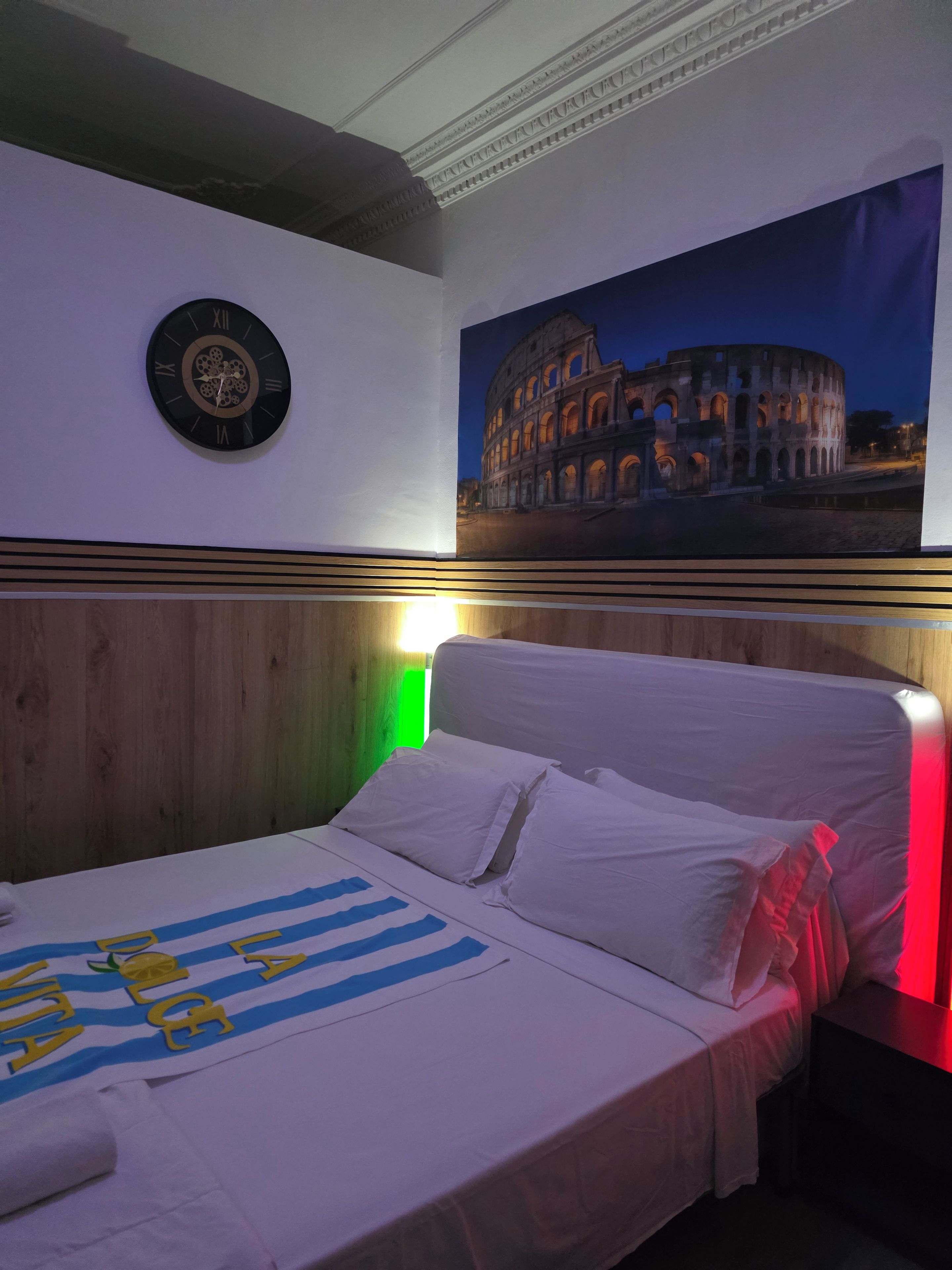 A bedroom with a bed, pillows, a clock, and a wall-mounted picture of the Colosseum.