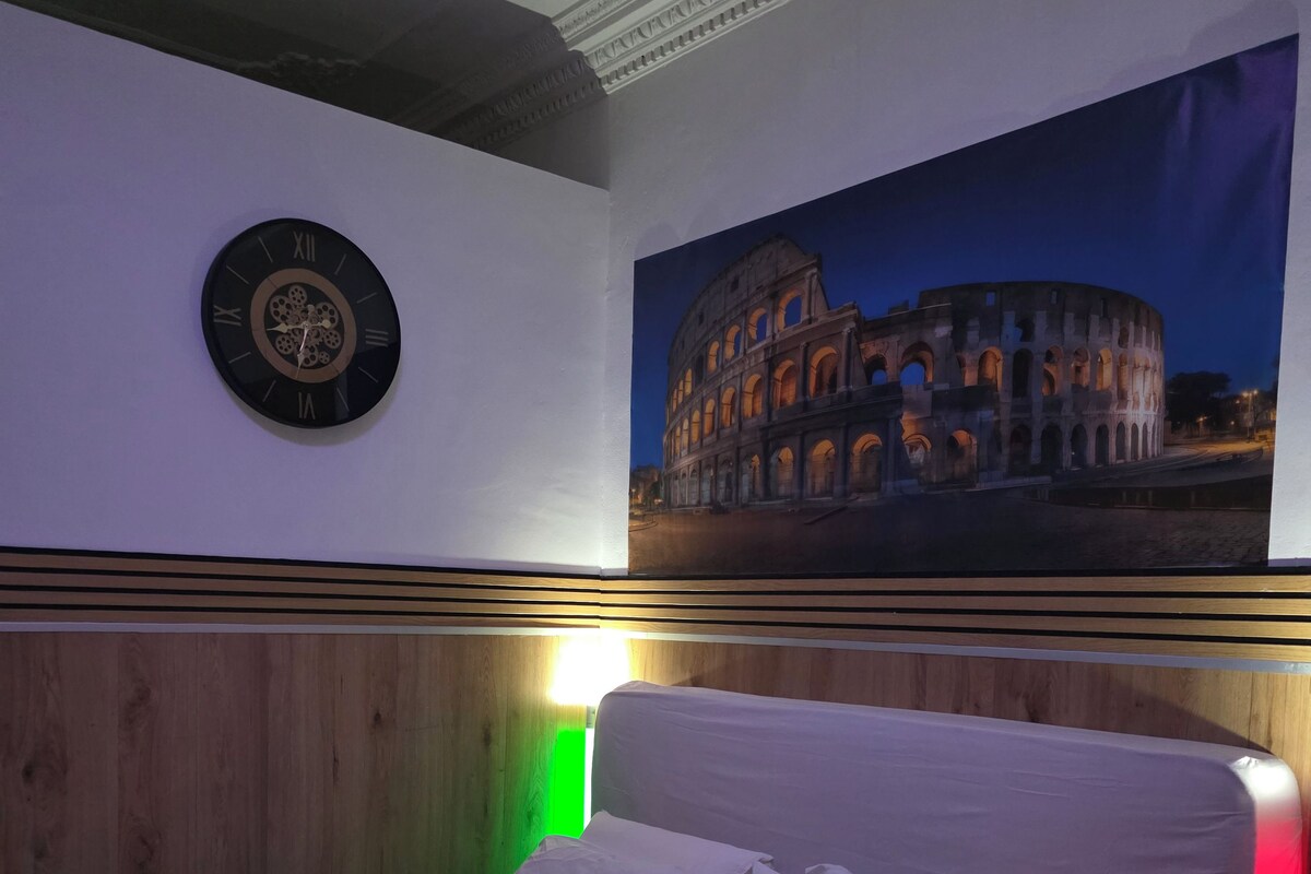 A bedroom with a bed, pillows, a clock, and a wall-mounted picture of the Colosseum.