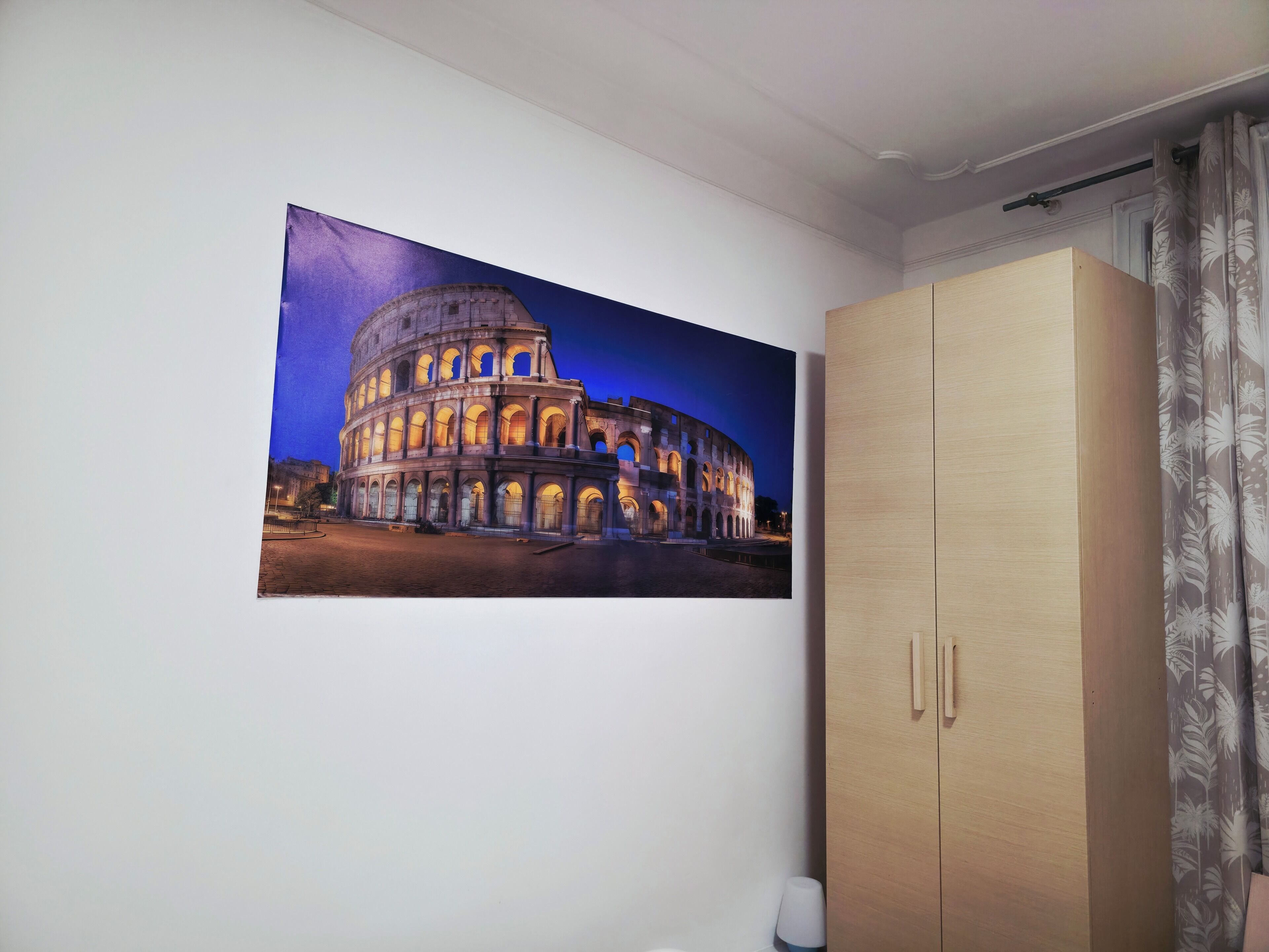 A room with a large framed picture of the Colosseum, a wooden wardrobe, and a curtain.