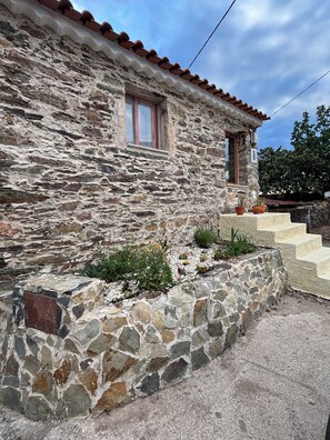 A stone building with a red-tiled roof, a window, and a small garden with plants and flowers.