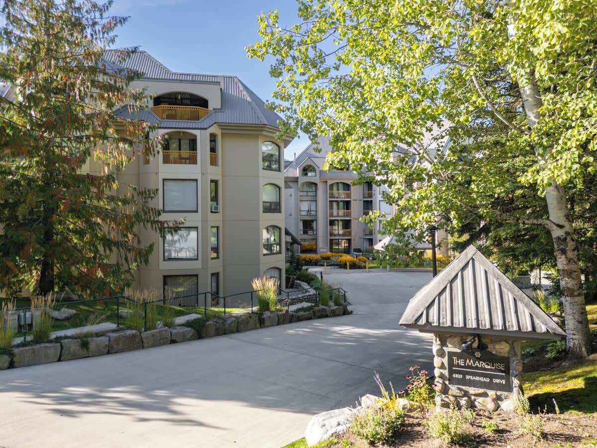 A multi-story building with a modern architectural style, a stone sign indicating the name 'The Marquise,' and a well-maintained landscaped area with trees and shrubs.