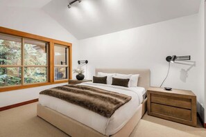 Een slaapkamer met een groot raam, een bed met een vachtdeken, een nachtkastje en een wandlamp.