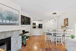 A modern kitchen with white cabinets, a dining area with white chairs, a stone fireplace, and a large painting on the wall. - Luxe 2-Bed | Ski-in Ski-out | On Hill (Whistler)