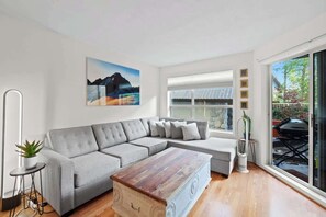 A modern living room with a grey sectional sofa, a wooden coffee table, and a large window with a view of the outdoors. - Luxe 2-Bed | Ski-in Ski-out | On Hill (Whistler)