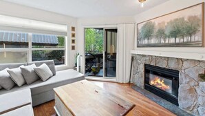 A living room with a stone fireplace, a sofa with cushions, a wooden coffee table, and a large window with a view of greenery outside. - Luxe 2-Bed Ski-in Ski-out, On Hill (Whistler)