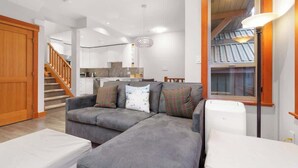 A modern living room with a grey sofa, a wooden door, a staircase, and a kitchen in the background. - Symphony - Walk to village & slopes - Luxury 4-bed (Whistler)