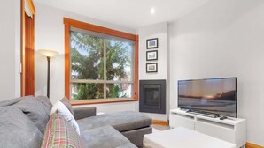 A modern living room with a grey sofa, a flat-screen TV, a fireplace, and a large window with a view of trees. - Symphony - Walk to village & slopes - Luxury 4-bed (Whistler)