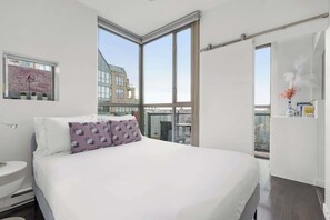 Ein modernes Hotelzimmer mit einem großen Bett, einem Nachttisch, einem Fenster mit Stadtblick und einem kleinen Fenster mit einer Pflanze.