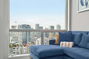 Ein blaues Sofa mit gestreifter Decke, ein Blick auf die Stadt durch ein großes Fenster und ein gerahmtes Kunstwerk an der Wand.