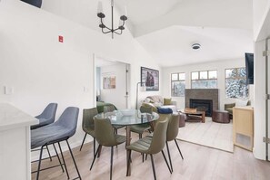 Un salon moderne avec une cheminée, un coin repas doté d’une table en verre et un espace cuisine.