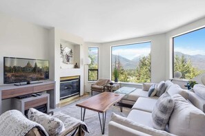 Ein modernes Wohnzimmer mit Kamin, Fernseher und großen Fenstern mit Blick auf die Berge.