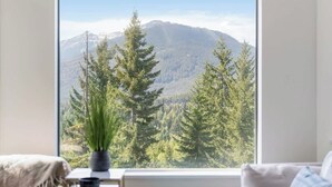Habitación con un amplio ventanal que ofrece vistas a la montaña, una planta en maceta sobre una mesa y una cama con cabecero blanco.