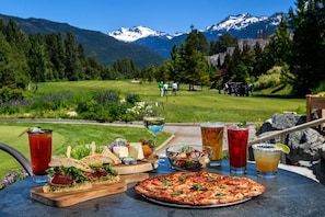 Ein Tisch mit Speisen und Getränken, eine gebirgige Landschaft und ein Golfplatz im Hintergrund.