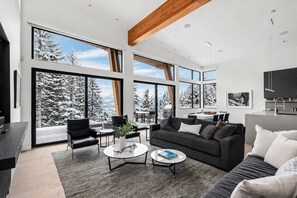 A modern living room with large windows, a dark sofa, a coffee table, and a dining area. - Timber Haven - Luxury Ski in Ski out - Kadenwood (Whistler)