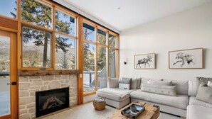 A modern living room with a stone fireplace, large windows, a grey sofa, and two framed animal prints on the wall. - Luxe 5-BED Scandinave Retreat - Walk to Slopes (Whistler)