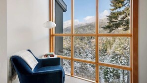 A room with a large window, a blue chair, and a small table with books. - Luxe 5-BED Scandinave Retreat - Walk to Slopes (Whistler)