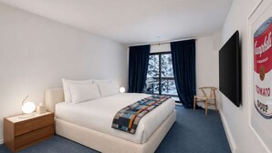 Una habitación de hotel con cama, una mesita de noche con lámpara, una silla, televisor y ventana con cortinas.
