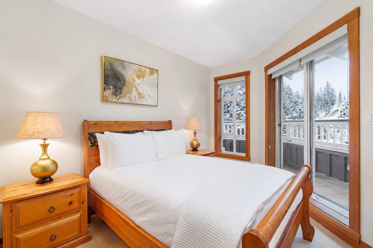 Une chambre à coucher avec un lit en bois, deux tables de chevet avec des lampes, une fenêtre donnant sur des arbres couverts de neige et un tableau accroché au mur.