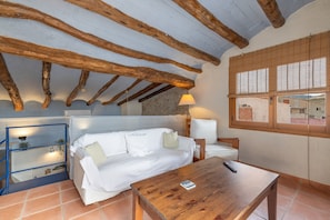A room with exposed wooden beams, a white sofa, a wooden table, a lamp, and a window with blinds. - Apartment 'Cal Capdevila Dúplex' with Mountain View, Wi-Fi and Air Conditioning (Pira)