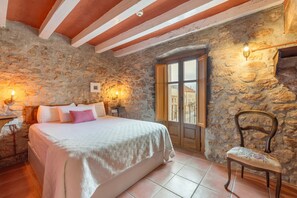 Une chambre avec un mur en pierre, un lit recouvert d’une courtepointe blanche, une chaise en bois et une fenêtre donnant sur un paysage agréable.