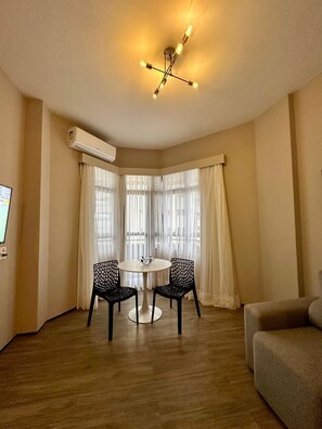 Quarto com mesa redonda, duas cadeiras, luminária no teto, ar condicionado e uma janela grande com cortinas.