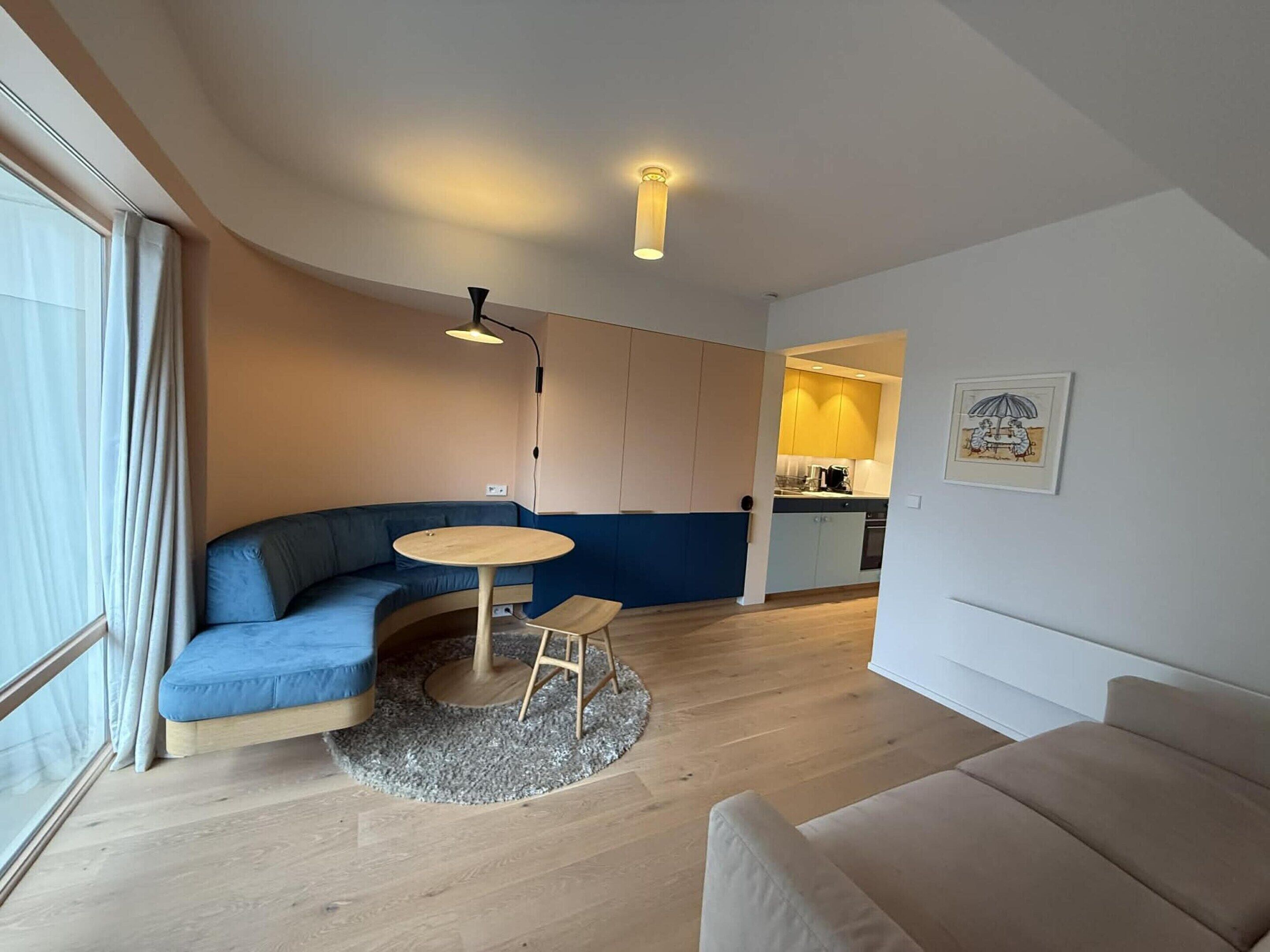 A modern living room with a blue sofa, a round coffee table, and a small stool. A kitchen is visible in the background with a yellow cabinet and a framed picture on the wall.
