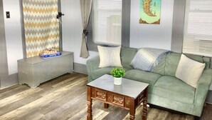 Ein Zimmer mit einem grünen Sofa, einem Holztisch, einem gemusterten Vorhang und einem Bild an der Wand.