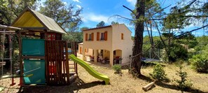 Une aire de jeux avec un toboggan et une petite structure en bois, entourée d’arbres et d’un bâtiment doté d’un toit en tuiles.