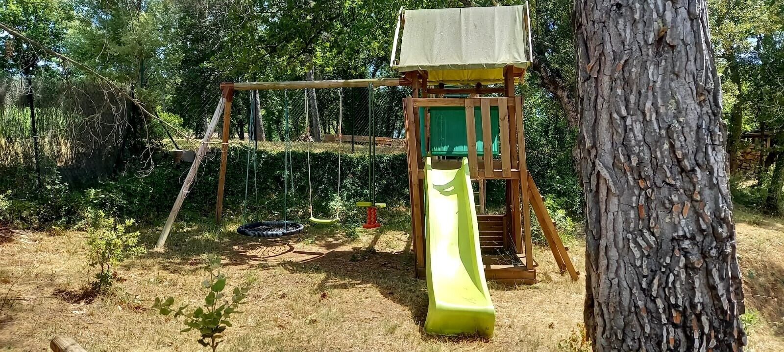 Un parc infantil amb tobogan i cadira de balanceig, envoltat d'arbres.