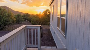 A wooden deck leading to a building with large windows, surrounded by trees and a mountain in the background.
