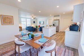 A modern kitchen with white cabinets, a central island, and a dining area with a wooden table and white chairs. - Grand Canyon Escape in Williams sleeps 6 (Williams)