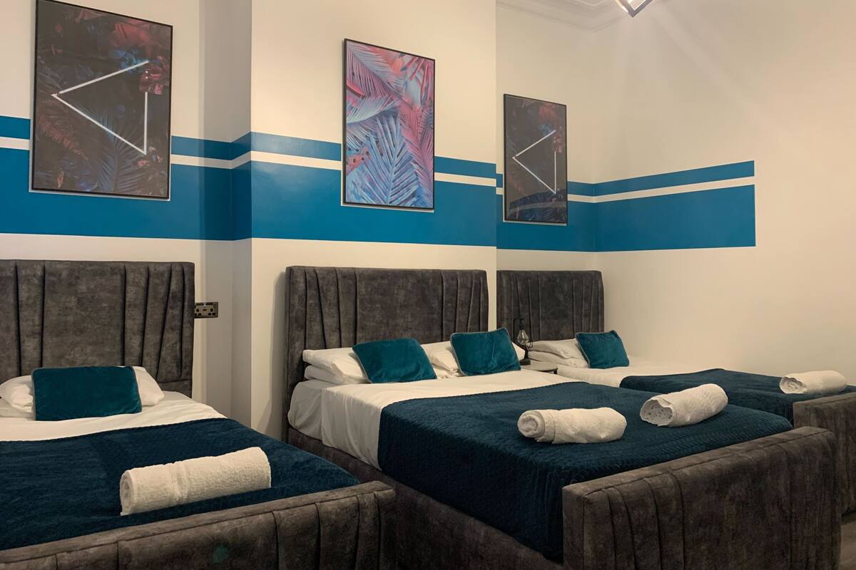A hotel room with three beds, each with a headboard, white linens, and teal pillows.