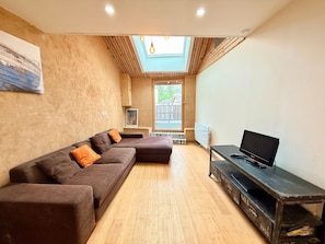 A modern living room with a sectional sofa, a television on a vintage cabinet, and a skylight above a balcony. - L’Authentique du Vercors – Spacious Modern Wood-Style Apartment with Mountain Views (Villard-de-Lans)